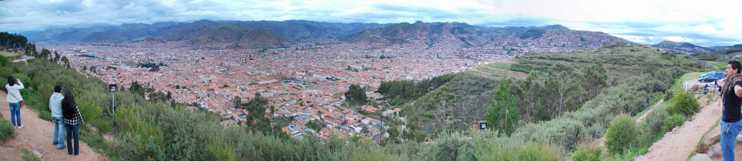 Panoramic view of Cusco, Peru from an elevated viewpoint showing dense cityscape with red terracotta rooftops filling a valley surrounded by Andes mountains under overcast sky, with tourists visible on dirt paths in the foreground