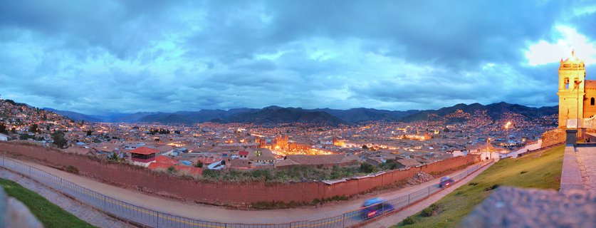 Panoramic cityscape of Arequipa Peru at blue hour showing illuminated colonial church tower on right, terracotta rooftops across valley, Andes mountains in background under cloudy twilight sky with light trails from vehicles on foreground road