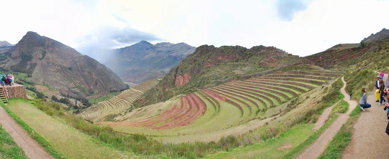 Panoramic view of ancient Incan agricultural terraces carved into a mountainside in Peru's Sacred Valley, with tourists on dirt paths and viewing platforms, mountains and cloudy sky in background