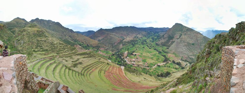 Panoramic view of curved Incan agricultural terraces carved into a green mountainside in Peru's Sacred Valley, with stone ruins in the foreground and mountain peaks under cloudy sky