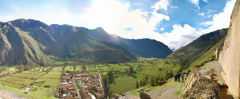Panoramic landscape showing ancient Incan stone ruins on a hillside overlooking a green valley with agricultural terraces, a small village, and towering Andes mountains under a blue sky with clouds