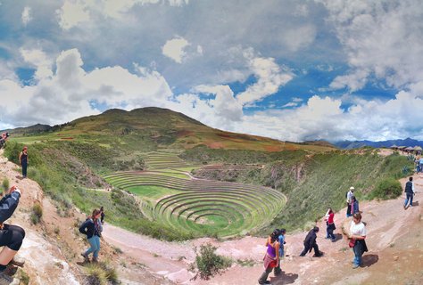 Aerial view of circular stepped agricultural terraces at Moray archaeological site with tourists on surrounding pathways, green mountains and blue cloudy sky in background