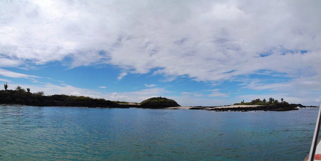 Panoramic coastal view of Galapagos Islands showing dark volcanic rock formations with sparse green vegetation rising from calm turquoise ocean water under blue sky with white clouds