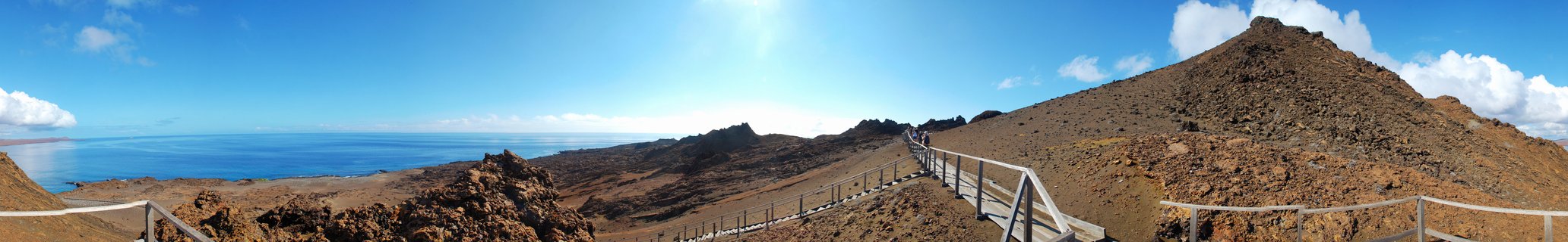 Wooden boardwalk with railings winding through brown volcanic rock terrain in Galápagos Islands, with ocean visible on the left and visitors walking on the path under blue sky
