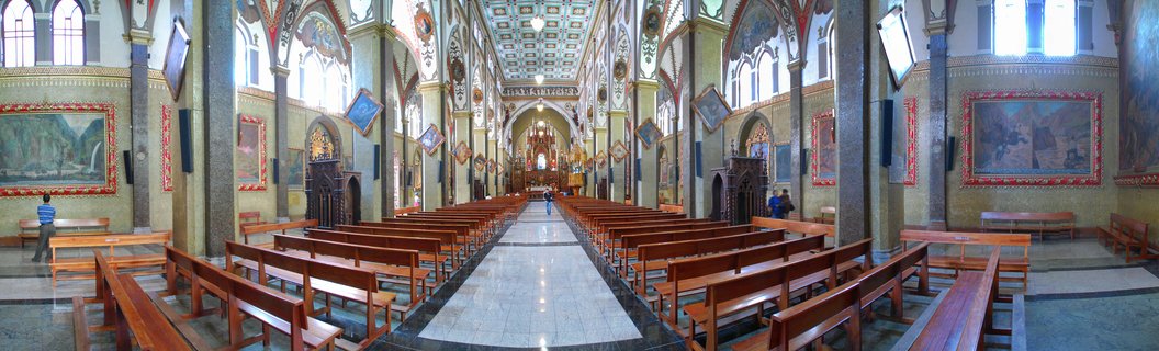 Panoramic view of a church interior showing rows of wooden pews on both sides of a central marble aisle, ornate decorated ceiling, large religious paintings on walls, stone columns, and altar at the far end
