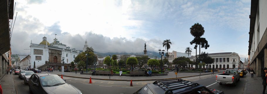 Wide street view of Plaza de la Independencia in Quito showing the white Presidential Palace with golden dome, colonial buildings, central monument, palm trees, pedestrians, and vehicles with Andes mountains in the background under cloudy sky