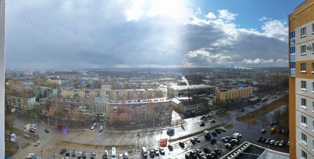 High-angle panoramic view of Russian city showing multi-story apartment buildings, wet streets with cars, industrial smokestacks, and cloudy sky with sun breaking through