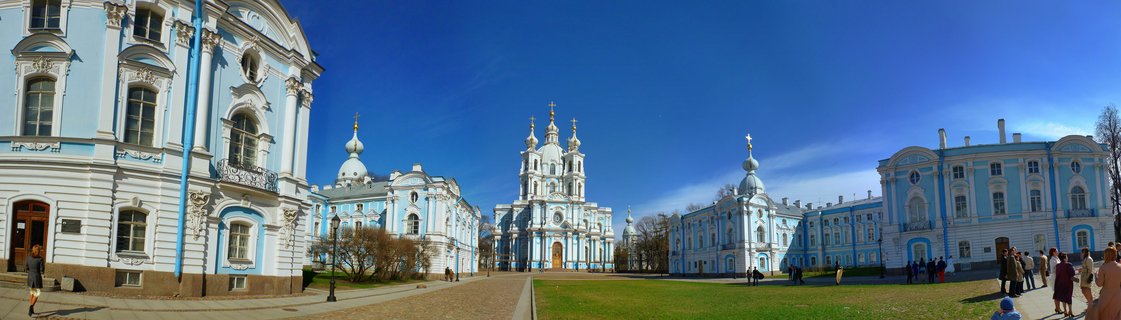 Panoramic view of the blue and white Smolny Cathedral complex with golden onion domes and Orthodox crosses in St. Petersburg, Russia on a sunny spring day with visitors walking on grounds