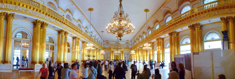 Interior view of a grand museum hall with golden columns, ornate chandeliers, white walls, and visitors walking on polished floors