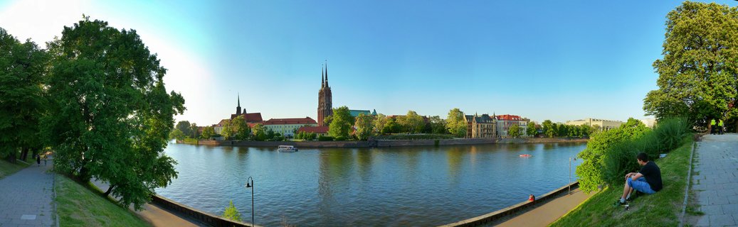 Panoramic view of the Cathedral of St. John the Baptist with twin Gothic spires on Ostrów Tumski in Wrocław, Poland, viewed across the Oder River during golden hour. The scene includes historic red-tiled rooftops, green spring trees along the riverbank, a person sitting on grass in the foreground right, walking paths along the river, a small boat on the water, and a clear blue sky with warm sunset lighting.