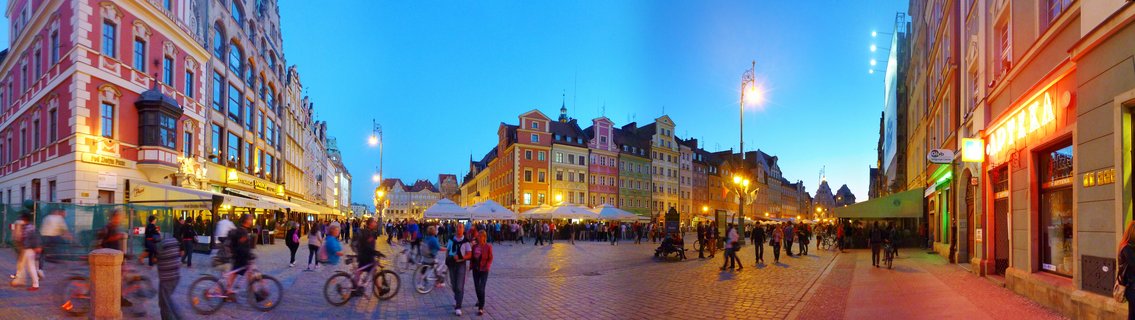 Wide view of Wrocław Market Square at twilight with colorful historic buildings, cobblestone pavement, pedestrians walking, cyclists riding, and illuminated street lamps under blue evening sky