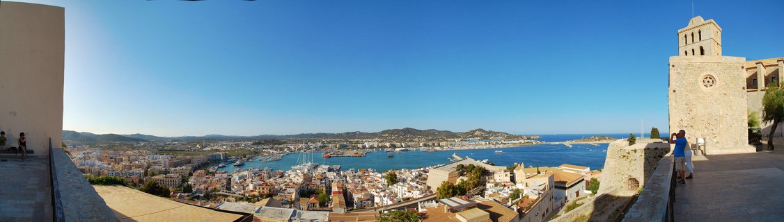 Panoramic view of Ibiza Town harbor from Dalt Vila old town, showing the Cathedral of Santa Maria d'Eivissa bell tower on the right, white Mediterranean buildings with terracotta roofs, sailboats and yachts in the blue harbor, and coastal mountains in the distance under a clear sky