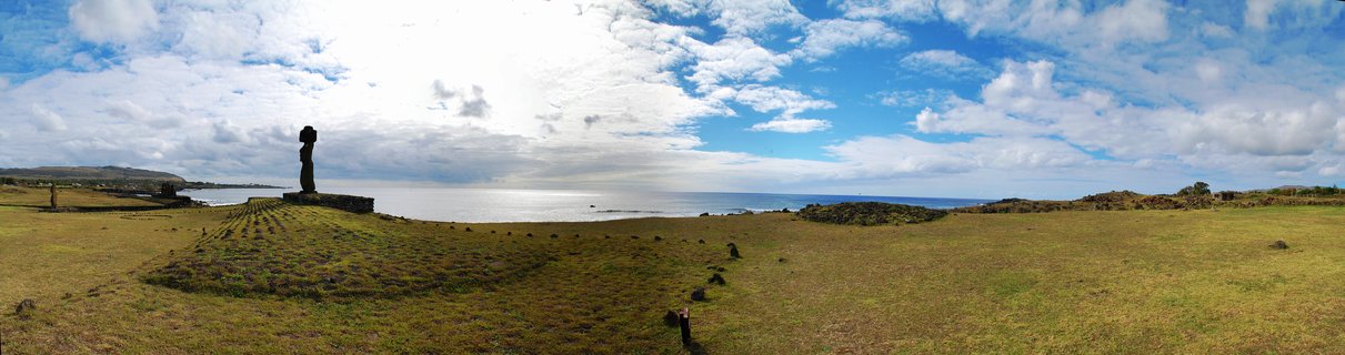 A large stone Moai statue standing on a raised grassy platform on Easter Island coastline with Pacific Ocean and cloudy blue sky in background