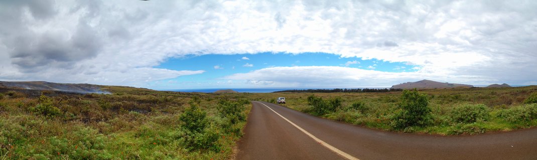 A curved paved road with yellow center line runs through green and brown grassy volcanic terrain on Easter Island, with a white vehicle, distant hills, ocean horizon, and cloudy sky with blue patches above.