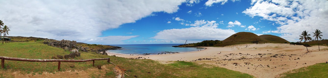 Wide panoramic coastal landscape showing a sandy beach curving along turquoise ocean water, with palm trees, green grass, volcanic hills, and blue sky with white clouds on Easter Island, Chile