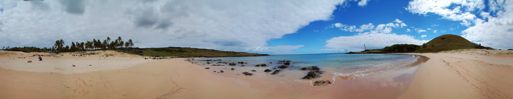 Wide panoramic beach scene showing golden sand, palm trees, calm blue ocean water with dark rocks, green hills, and cloudy sky on Easter Island
