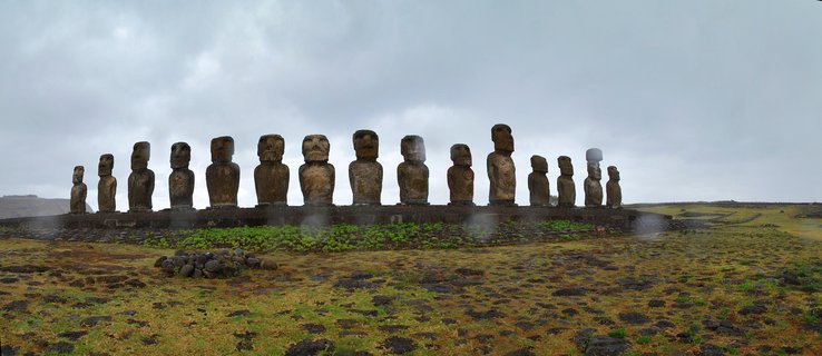 Fifteen large stone moai statues standing in a row on a raised stone platform at Ahu Tongariki archaeological site on Easter Island, Chile, with green grass in the foreground and overcast sky above
