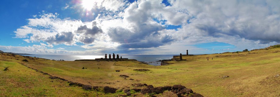 Panoramic view of ancient Moai stone statues on a ceremonial platform at Easter Island, with grassy terrain, Pacific Ocean, and cloudy sky with sun visible