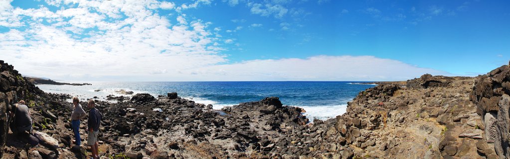 Panoramic view of three people standing on black volcanic rocks along Easter Island's coastline with blue Pacific Ocean and cloudy sky