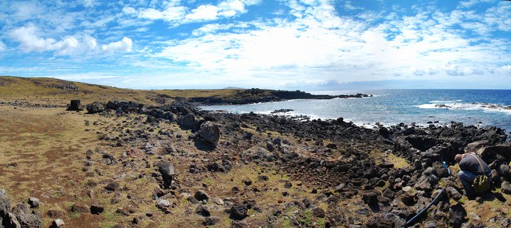Panoramic view of rocky volcanic coastline on Easter Island with scattered basalt boulders, dry grassland, Pacific Ocean waves, blue sky with clouds, and a photographer in the foreground