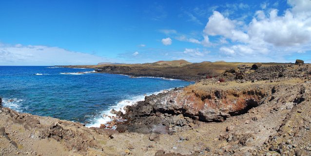 Panoramic view of rocky volcanic coastline with turquoise ocean water, white waves, brown and reddish lava rocks in foreground, distant hills under blue sky with white clouds