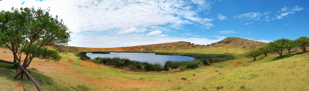 Panoramic view of a volcanic crater lake surrounded by green grassland, with trees on both sides and hills in the background under a blue sky with white clouds