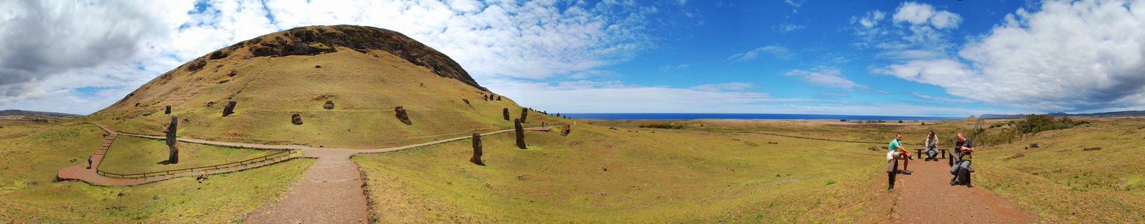 Panoramic view of Moai stone statues on grassy volcanic slopes at Rano Raraku quarry, Easter Island, with tourists on pathways and ocean visible in the distance under blue sky with clouds.