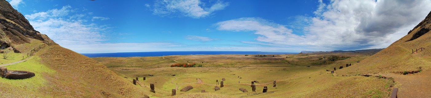 Panoramic view of Rano Raraku volcanic crater showing multiple Moai statues scattered across grassy slopes with Pacific Ocean horizon and blue sky with clouds
