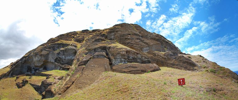 Volcanic rock quarry with grassy slopes showing partially carved stone moai statue embedded in the cliff face under blue sky with white clouds, Easter Island, Chile