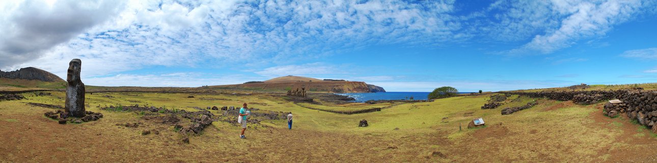 Panoramic landscape showing ancient Moai stone statues on Easter Island with tourists walking on grassy terrain, volcanic stone walls, ocean coastline, and blue sky with clouds