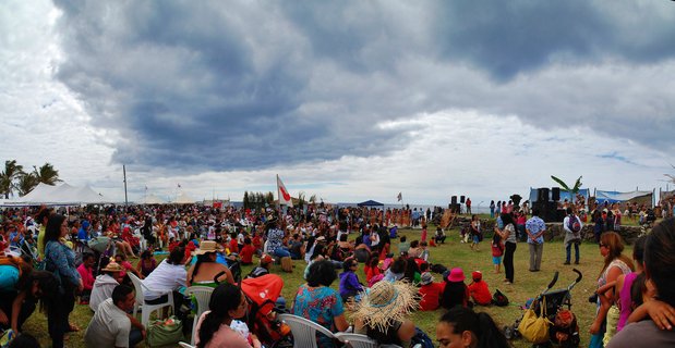 Large outdoor crowd seated and standing on grassy field near ocean under cloudy sky at Easter Island festival with white tents and stage equipment visible