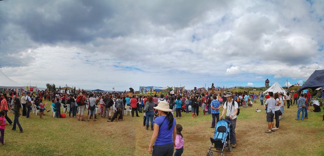 Wide panoramic view of a large outdoor cultural festival on Easter Island with hundreds of people on a grassy field, white tents, and a moai statue visible on a hill in the background under cloudy skies