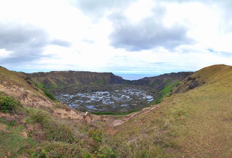 Aerial view looking down into Rano Kau volcanic crater on Easter Island, showing a marshy lake with vegetation patches, surrounded by grassy crater slopes under cloudy skies with ocean visible in the distance