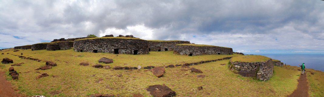 Panoramic view of ancient stone structure ruins on a grassy hillside overlooking the ocean under cloudy sky at Easter Island archaeological site