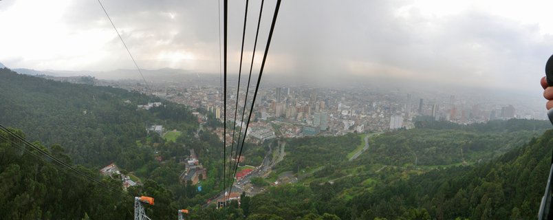 Panoramic aerial photograph of Bogotá city viewed from cable car, with multiple black cables in foreground, green forested mountains, and urban buildings stretching to distant hills under cloudy sky