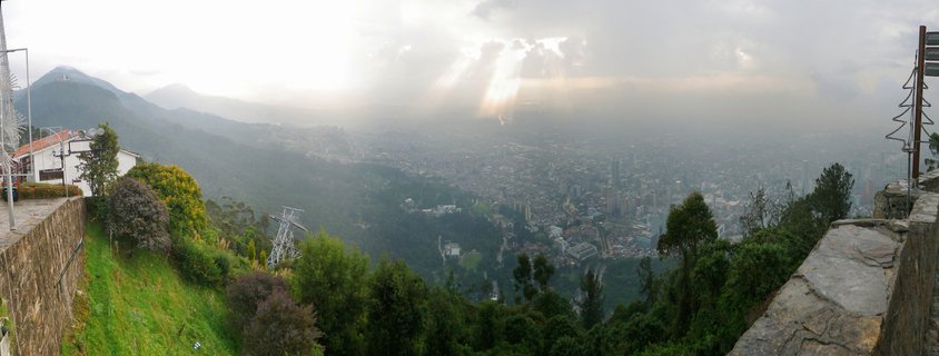 Panoramic cityscape of Bogotá, Colombia showing urban sprawl in a valley surrounded by Andes mountains, with dramatic sun rays breaking through clouds, viewed from an elevated stone viewpoint with vegetation in foreground