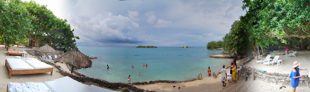 Panoramic beach scene showing a tropical resort in Cartagena with turquoise Caribbean water, people swimming, thatched umbrellas, white lounge chairs, and green trees on both sides of a curved bay