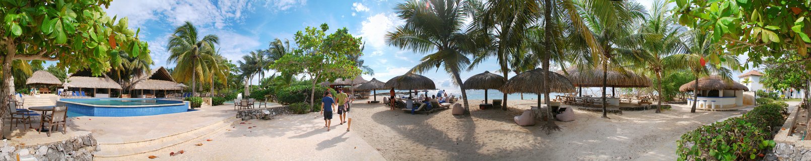 Panoramic view of a tropical beach resort showing a blue swimming pool on the left, thatched umbrellas and palm trees across sandy grounds, with people walking toward the ocean in Cartagena, Colombia.
