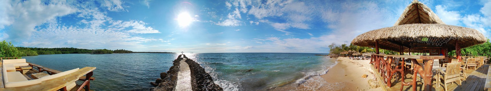 Panoramic beach scene showing a thatched-roof palapa restaurant on sandy shore, stone pier extending into turquoise Caribbean water, wooden deck furniture, palm trees, and blue sky with white clouds in Cartagena, Colombia