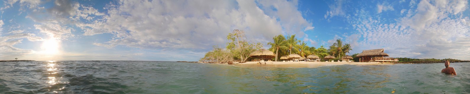 Panoramic view of tropical beach with thatched-roof bungalows, palm trees, and a person swimming in turquoise Caribbean waters at sunset in Cartagena, Colombia