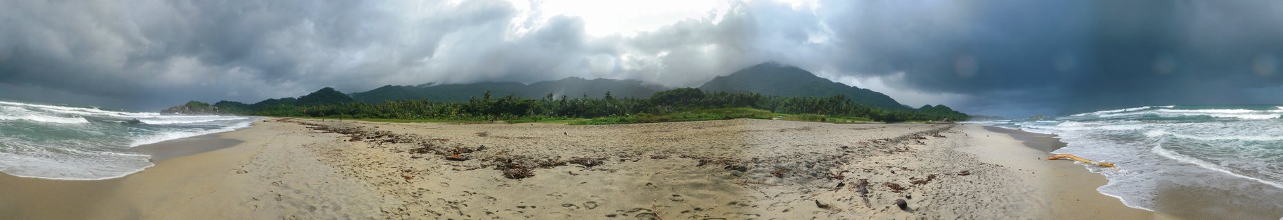 Wide panoramic beach scene showing sandy shore with seaweed, ocean waves on both sides, palm trees, mountains in background under dramatic stormy clouds