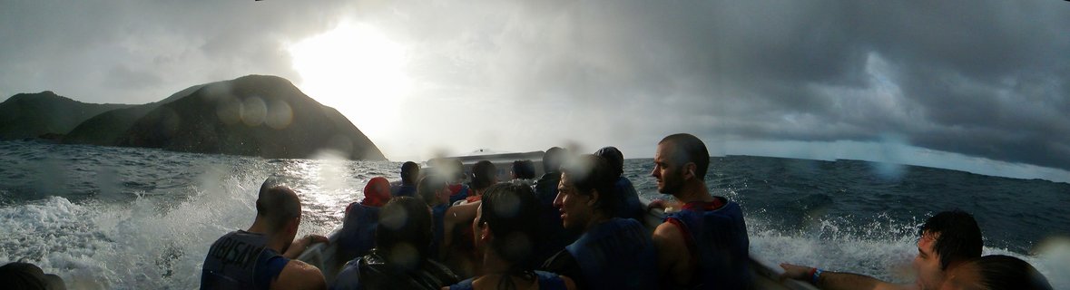 Group of tourists in blue life vests on a moving boat with white water spray, dark storm clouds overhead with bright sun breaking through, mountainous island visible in the background on the Caribbean Sea near Santa Marta, Colombia