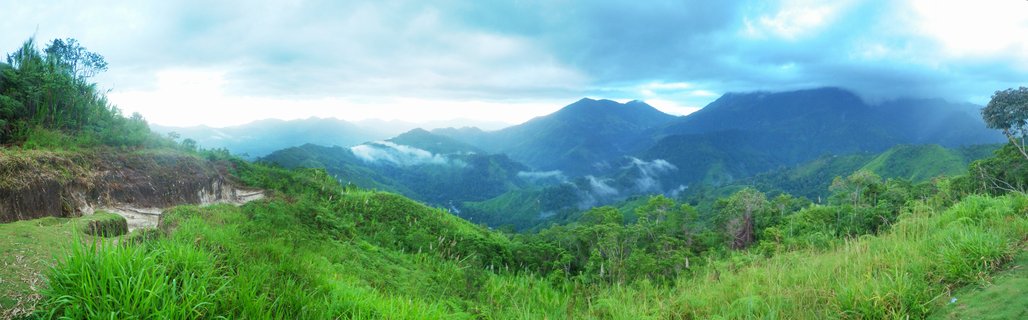Wide mountain landscape showing green tropical vegetation covering rolling hills and valleys with mist in the distance under cloudy sky
