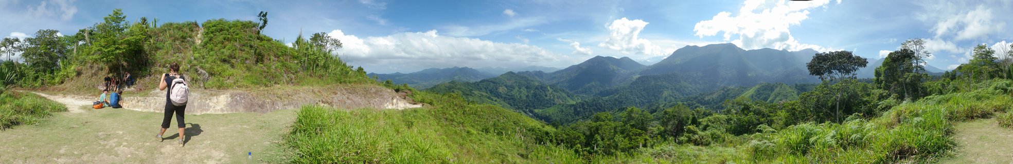 A person with hiking backpack stands on a dirt path viewpoint overlooking green mountain ranges and tropical forest under blue sky with white clouds in Colombia