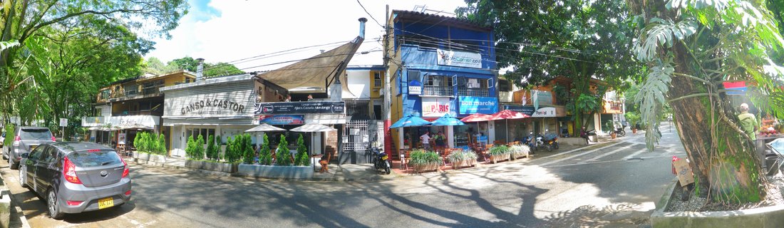 Street view showing restaurants with outdoor seating, colorful umbrellas, parked cars, shops with signs, tropical trees, and buildings in Medellín, Colombia