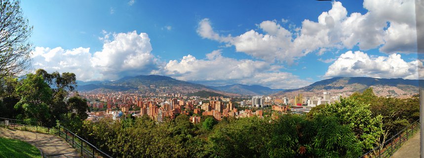 Panoramic cityscape of Medellin Colombia showing dense urban buildings in a mountain valley with green vegetation in foreground and blue sky with white clouds