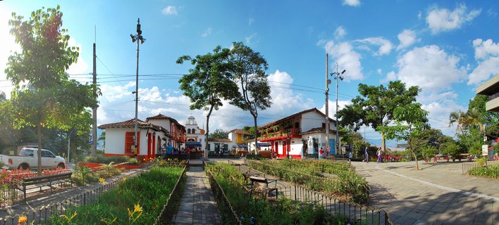 Panoramic view of a Colombian colonial town square with white buildings featuring red trim, a church bell tower, trees, benches, walkways, and people walking under a blue sky with clouds
