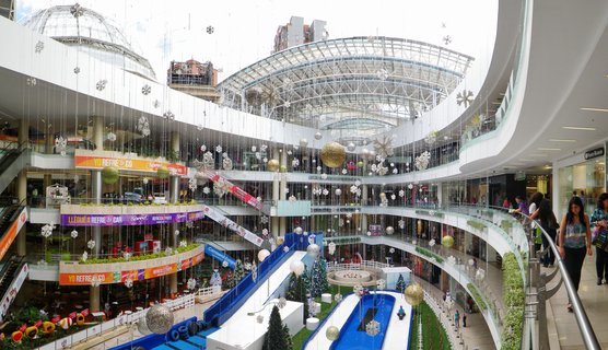 Interior view of a multi-level shopping mall with curved white balconies, glass roof, hanging snowflake and sphere holiday decorations, Christmas trees, blue slide feature, and shoppers on multiple floors