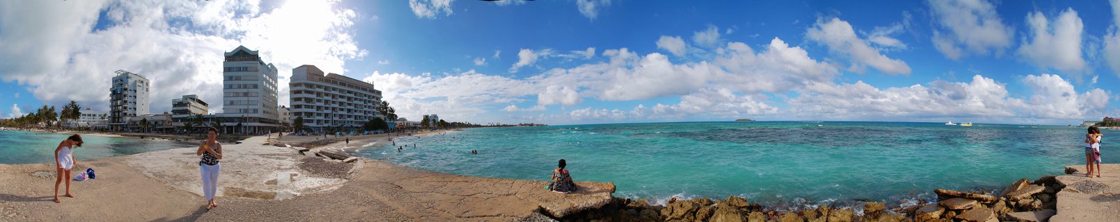 Panoramic view of San Andres Island beach in Colombia showing turquoise Caribbean water, white hotel buildings, tourists on sandy beach and rocky seawall under blue sky with clouds.