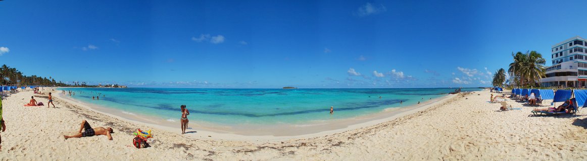 Wide panoramic beach scene showing white sand, turquoise ocean water, people sunbathing and swimming, palm trees on the left, hotel buildings and blue cabanas on the right, clear blue sky above
